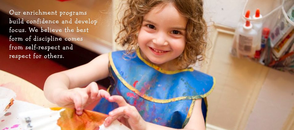 Our enrichment programs build confidence and develop focus. A smiling girl in a painting smock does an art project.